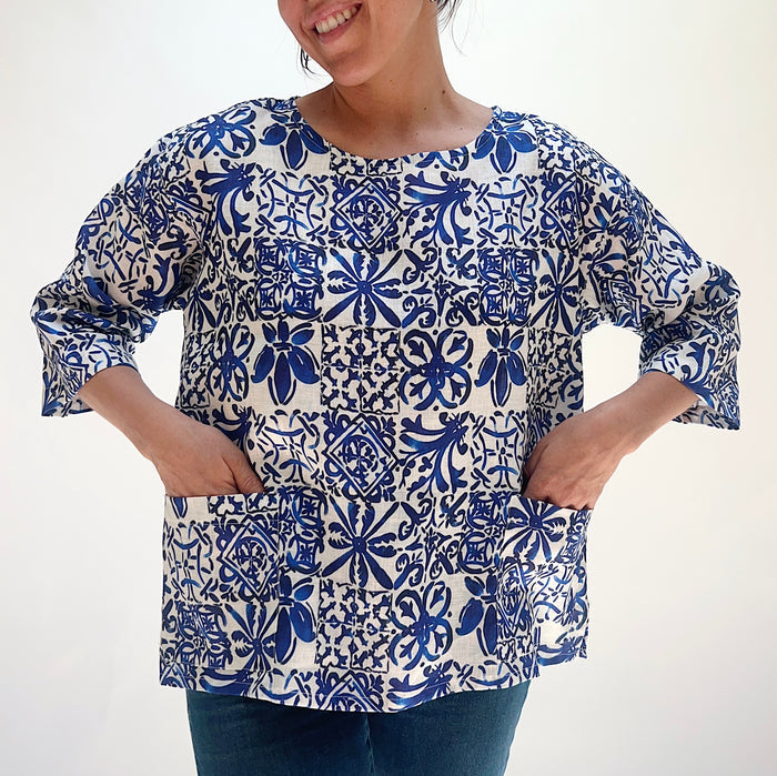 Front view of the Christopher Calvin Linen Blue Tiles Print Top with hands in the two front patch pockets, showing the relaxed silhouette, boat neckline, and blue-and-white tile print, on a white background