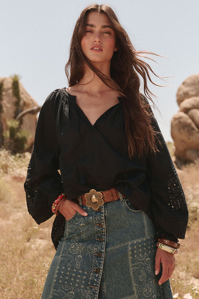 Front view of black schiffli anglaise blouse with blouson sleeves, ball-trim neckline, styled with denim skirt outdoors