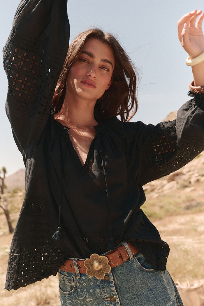Front view of black blouse with broderie anglaise sleeves, tassel ties, and flouson cuffs, styled with denim