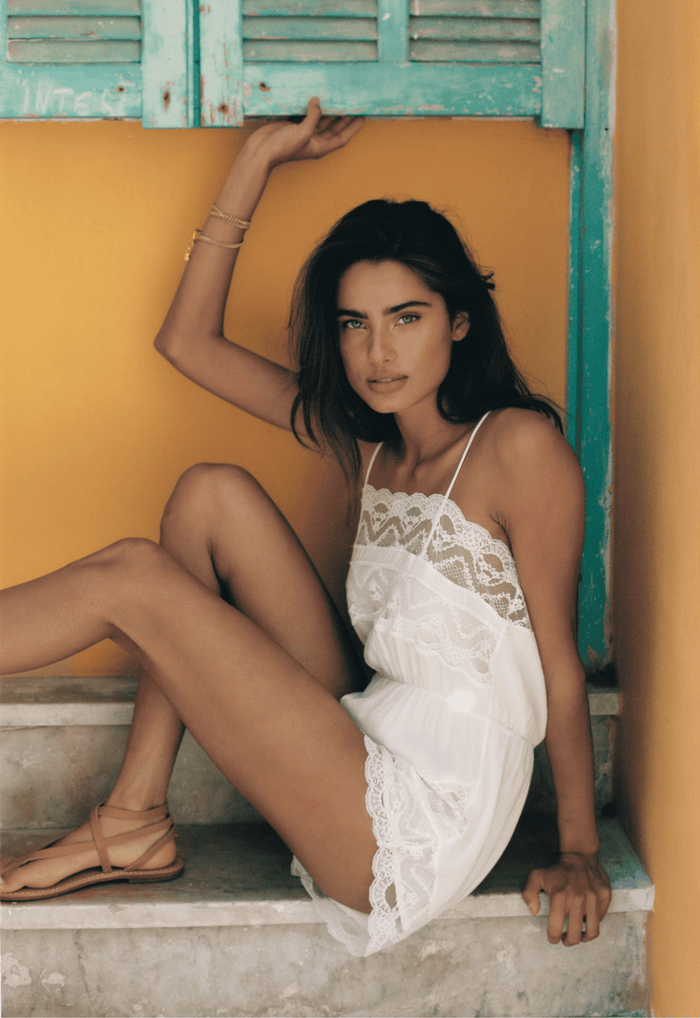 Model seated front-facing in ivory Mila Lace Romper, showing scallop lace neckline and adjustable straps