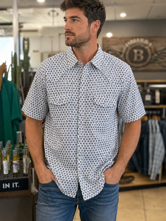 A man wearing a short-sleeve button-down shirt with a white and navy blue geometric pattern, paired with blue jeans, standing inside a retail store.