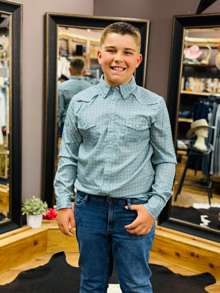 A young boy smiling in a retail store, wearing a light blue and white patterned western-style button-down shirt with blue jeans.