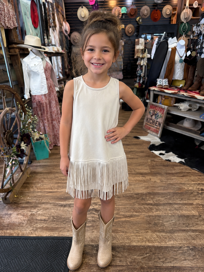 Young girl in a beige fringe dress and boots standing in a store with hats and clothing in the background.