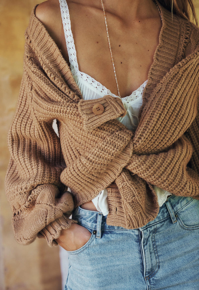 Model styled open with arms crossed, showing blouson sleeve, cable knit detail and single button cuff in toasted almond