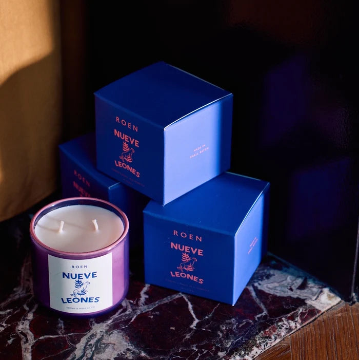A Roen candle with a label 'NUEVE LEONES' displayed on a marble surface with blue product boxes stacked next to it.