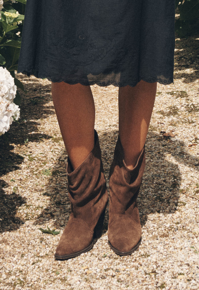 Close-up of chocolate suede slouch boots with wood stack heel, styled with black embroidered midi skirt on gravel