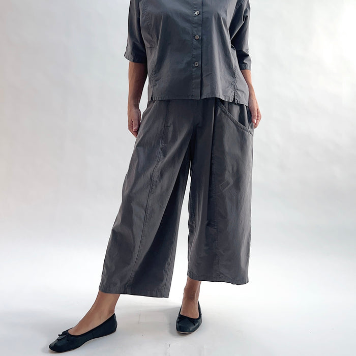 Full front view of model wearing Eleven Stitch eiffel tower one pocket pant with hands in pockets, showing elastic waistband, wide curved-seam cropped leg, large side pocket, gathered elastic hem, and deep slate grey color, styled with matching grey short sleeve button front shirt and black ballet flats