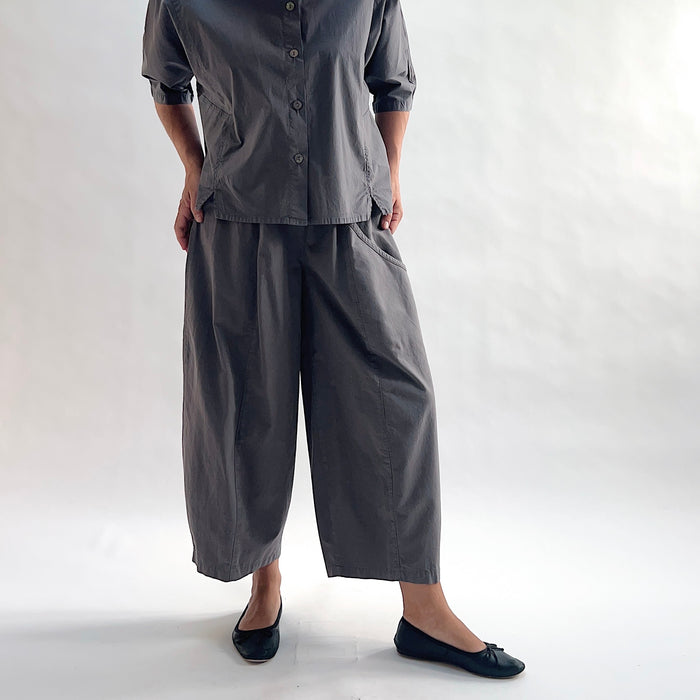 Close front view of model wearing Eleven Stitch eiffel tower one pocket pant with hands at sides, showing elastic waistband, wide curved-seam cropped leg, large curved side pocket, and deep slate grey color, styled with matching grey short sleeve button front shirt and black ballet flats