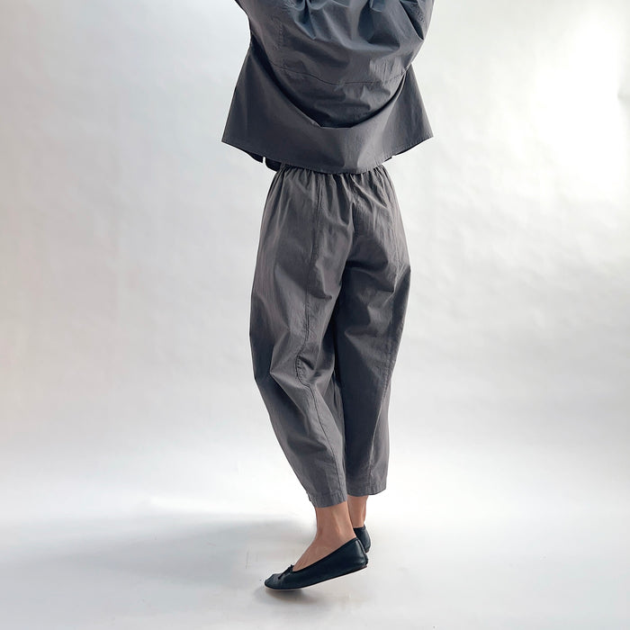 Three-quarter side view of model wearing Eleven Stitch eiffel tower one pocket pant, showing elastic waistband, wide curved-seam cropped leg, large side pocket, gathered elastic hem, and deep slate grey color, styled with matching grey short sleeve button front shirt and black ballet flats