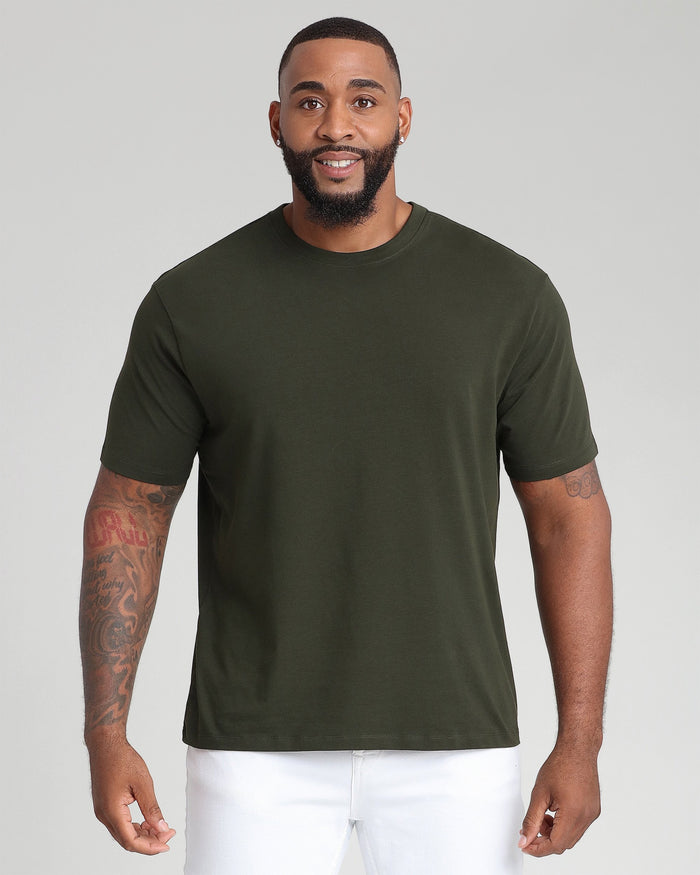 Man wearing olive green crew neck t-shirt with white pants against gray background.