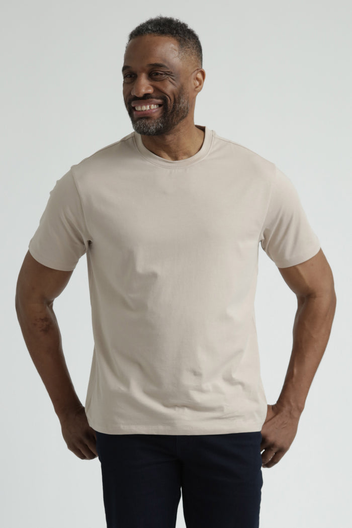 Man wearing tan crew neck t-shirt with short sleeves and navy pants against white background.