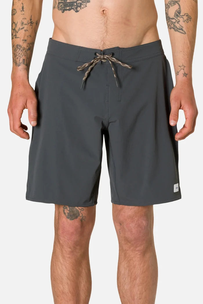 Front view of a tattooed man wearing the Katin OTG Crest Surf Trunk in Black Wash, showing the dark charcoal grey board short with braided drawstring and Katin label, against a white background