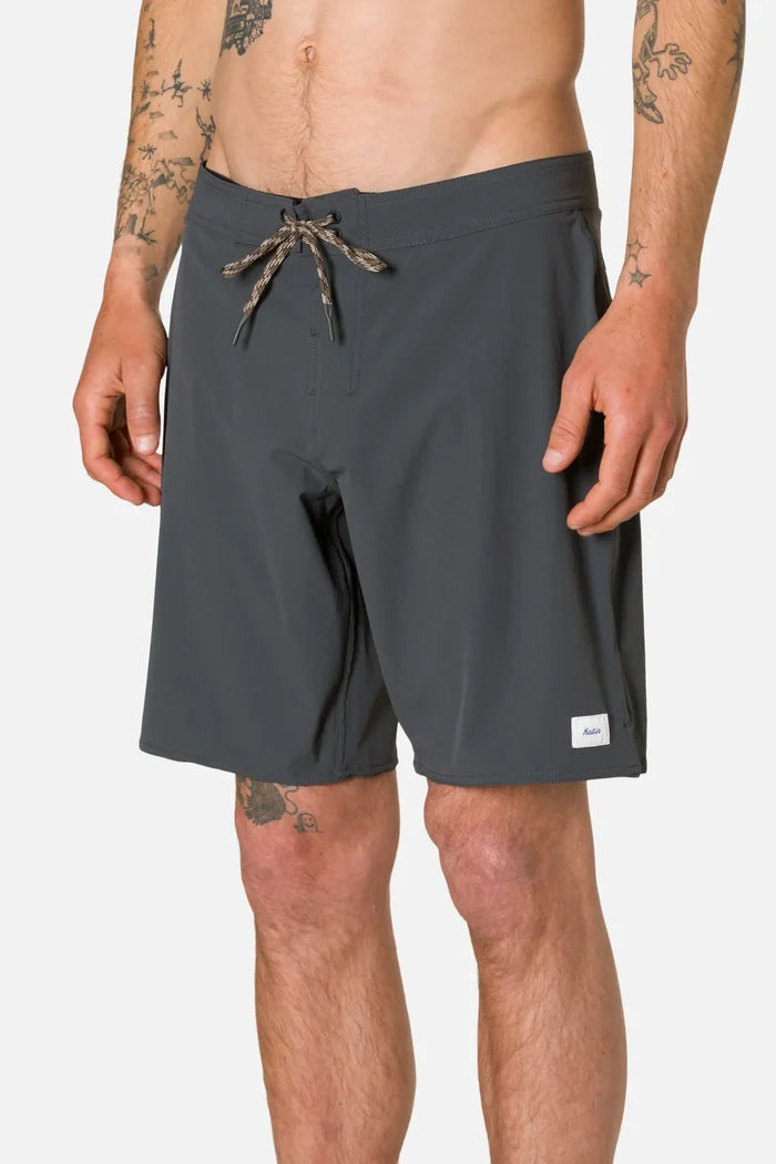 Three-quarter front view of a tattooed man wearing the Katin OTG Crest Surf Trunk in Black Wash, showing the side seam, spandex gusset fly closure, and Katin label on the lower right leg