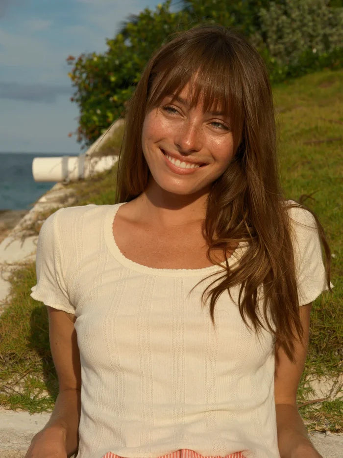 Woman with long brown hair smiling and wearing a cream colored pointelle tee with cap sleeves. She sits outdoors near a body of water.