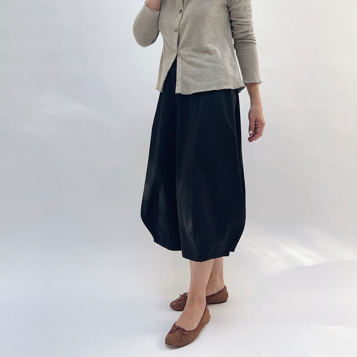 Rib Yoke Skirt in Black, side view, below-the-knee length with sculptural hem, styled with a beige draped top and brown flats