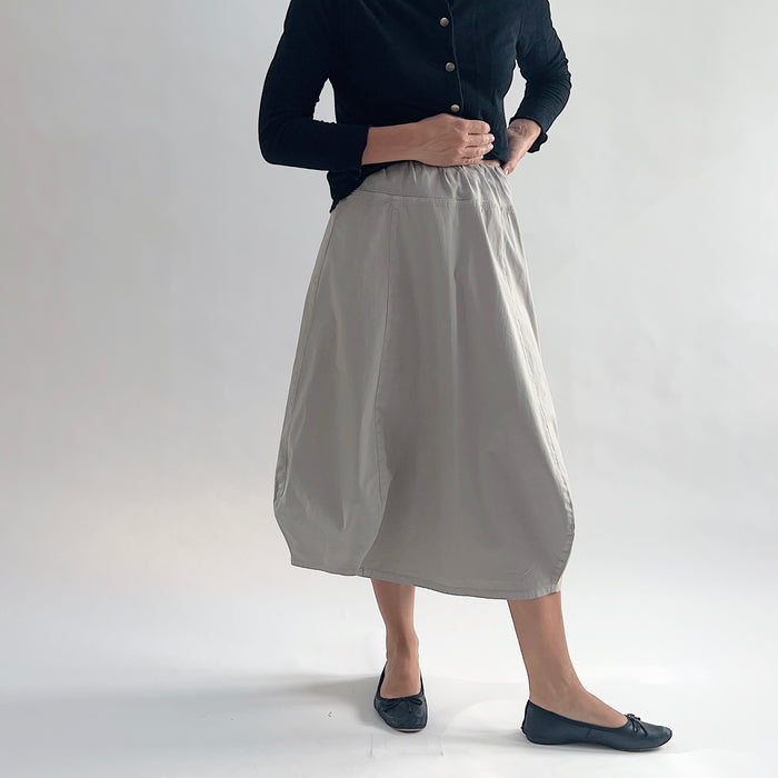Full front view of model wearing Eleven Stitch fennel rib yoke skirt with hands on hips, showing elastic knit waistband, curved seam paneling, sculptural bubble hem, below-the-knee length, and warm taupe color, styled with black button-front cardigan and black ballet flats