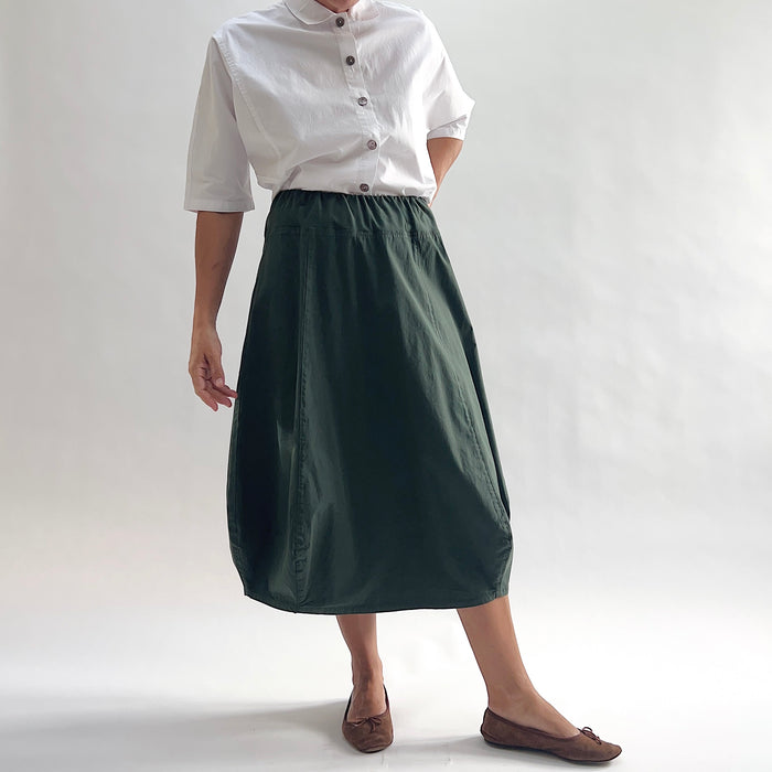 Full front view of model wearing Eleven Stitch konbu rib yoke skirt with arm at side, showing elastic knit waistband, curved seam paneling, sculptural bubble hem, below-the-knee length, and deep forest green color, styled with white chalk camp shirt and brown ballet flats