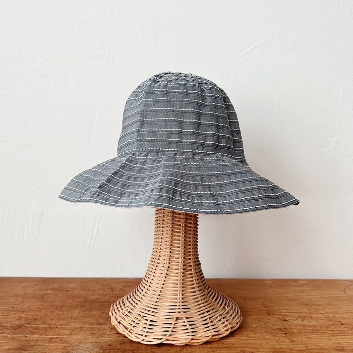 Sage ribbon bucket hat with white stitch stripe detail and wide brim, shown on a wicker hat stand — front view