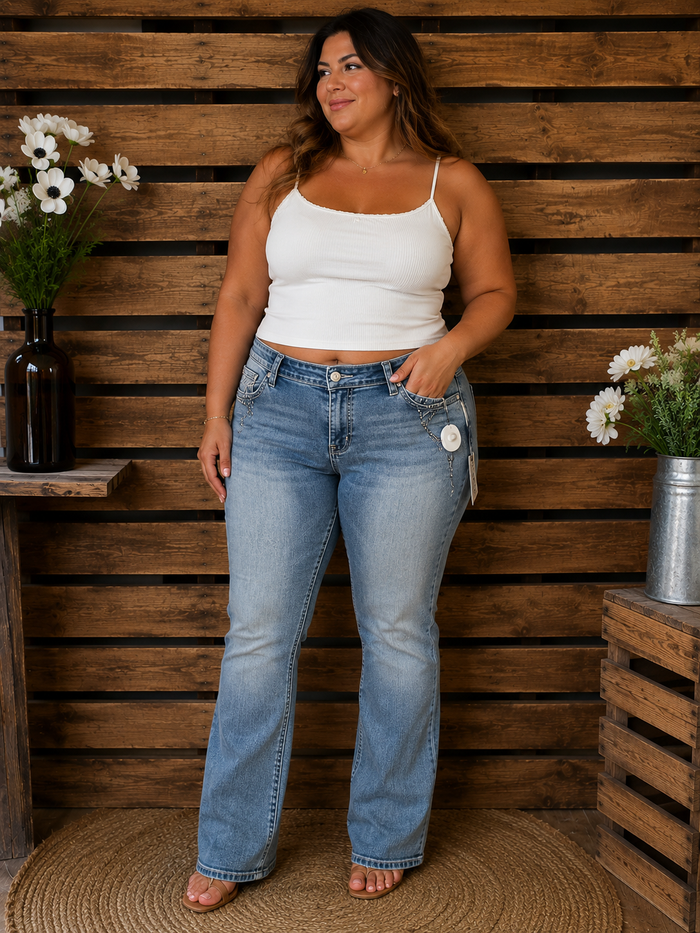 Woman wearing a white top and blue jeans standing against a wooden wall with plants.