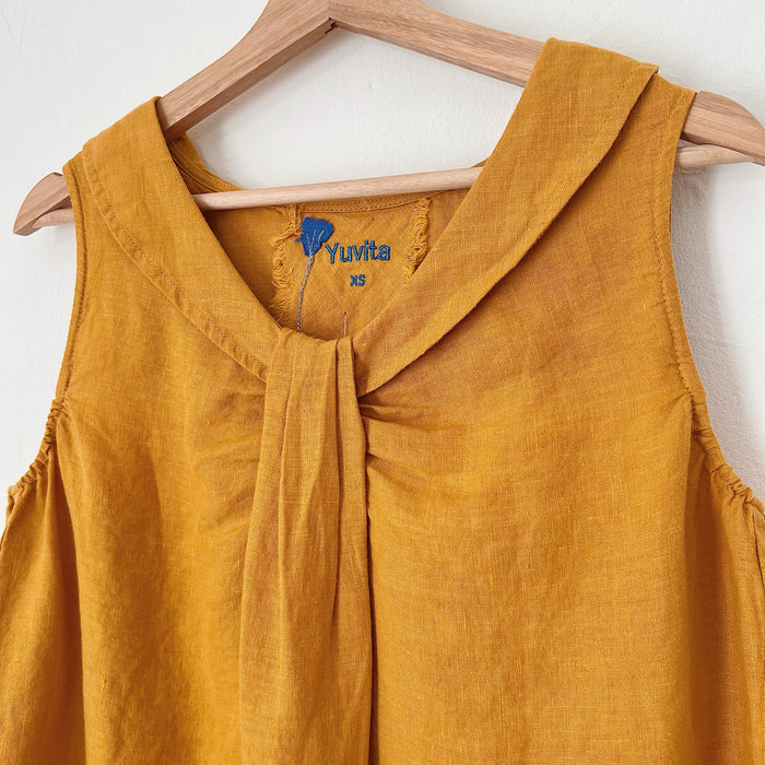 A close up shot of a sleeveless, turmeric yellow linen top hanging on a wooden hanger against a white background. The tag says the name of the brand, Yuvita.