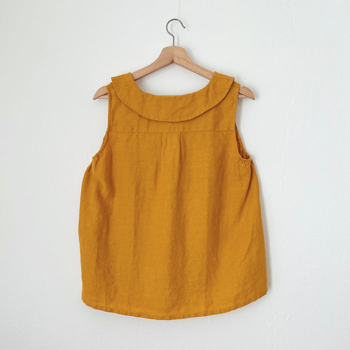 A sleeveless, turmeric yellow linen top hanging on a wooden hanger against a white background as seen from the back of the tank.