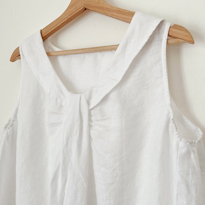 A close up shot of a white linen sleeveless top with a round neckline, hanging on a wooden hanger against a white wall.