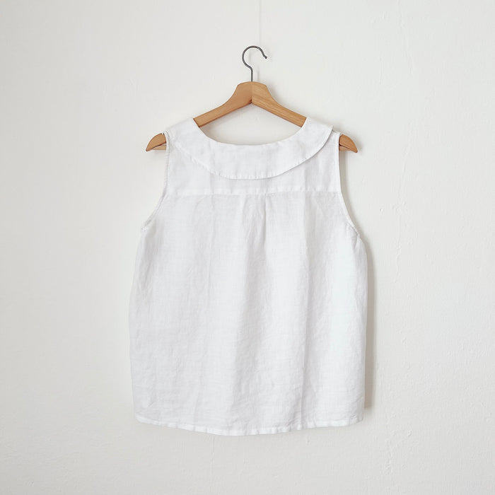 A view of the back of a white linen sleeveless top with a round neckline, hanging on a wooden hanger against a white wall.