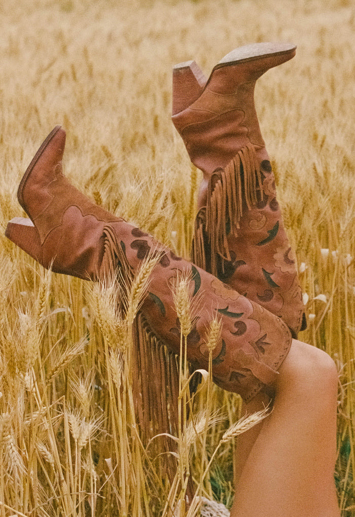 Legs raised in wheat field wearing tan suede fringe boots with intricate floral inlay and contrast tassels