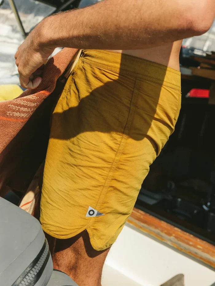 Mollusk Surf Shop Scallop Trunks in Mustard, close-up side view showing Velcro fly, back patch pocket, and pennant patch logo, worn on a boat in sunlight