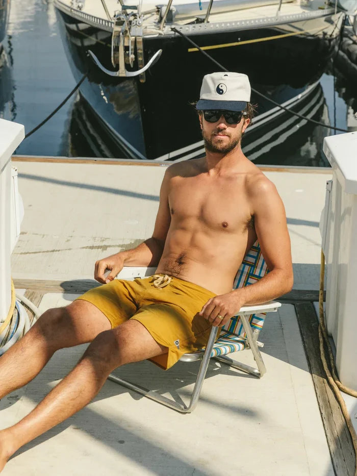 Image 4 Man wearing Mollusk Surf Shop Scallop Trunks in Mustard relaxing in a folding chair on a boat deck at a marina, wearing sunglasses and a yin-yang cap
