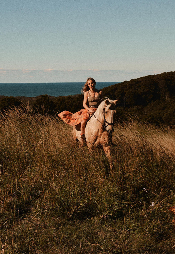 A woman rides a light-colored horse through tall dry grass on a hillside, with ocean and dark shrubbery in the background under a pale blue sky.
