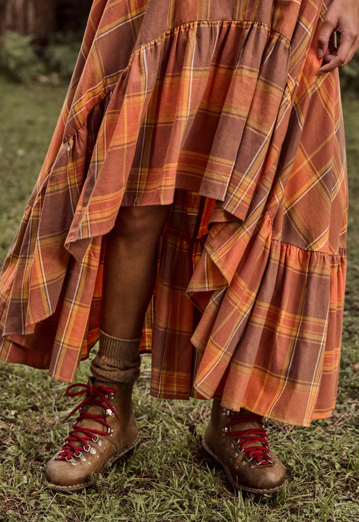 A close-up outdoor shot of a person wearing a long tiered plaid skirt in rust orange, brown, and yellow tones, paired with brown lace-up hiking boots on grass.