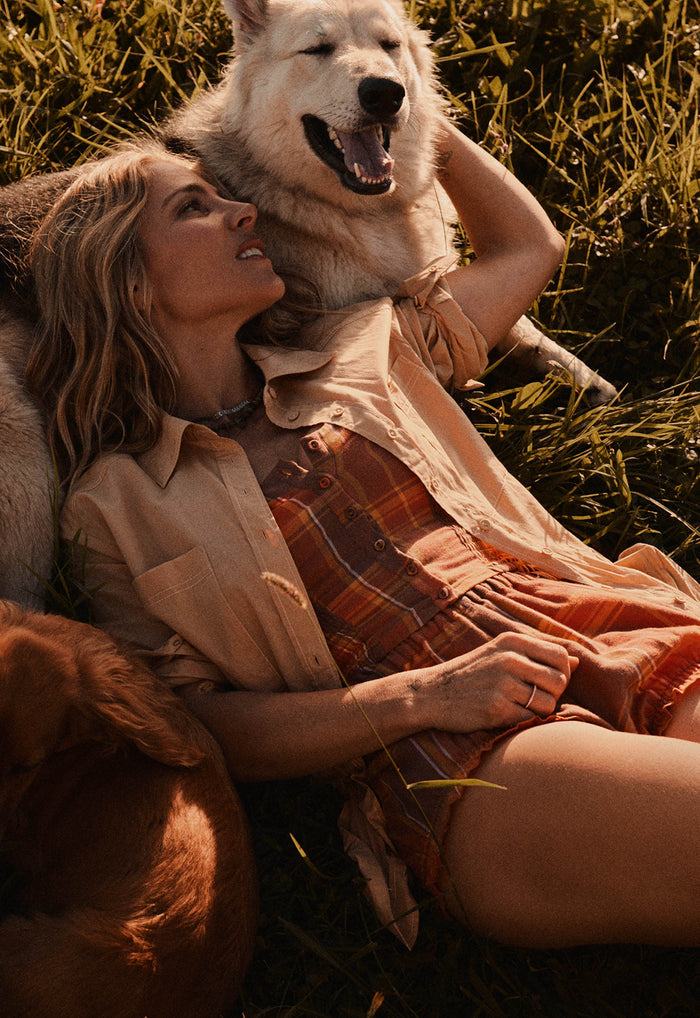 A woman lying in grass outdoors with a large white dog behind her and a brown dog at her side, wearing a beige shirt and rust-orange plaid shorts or dress. Warm golden sunlight illuminates the scene.