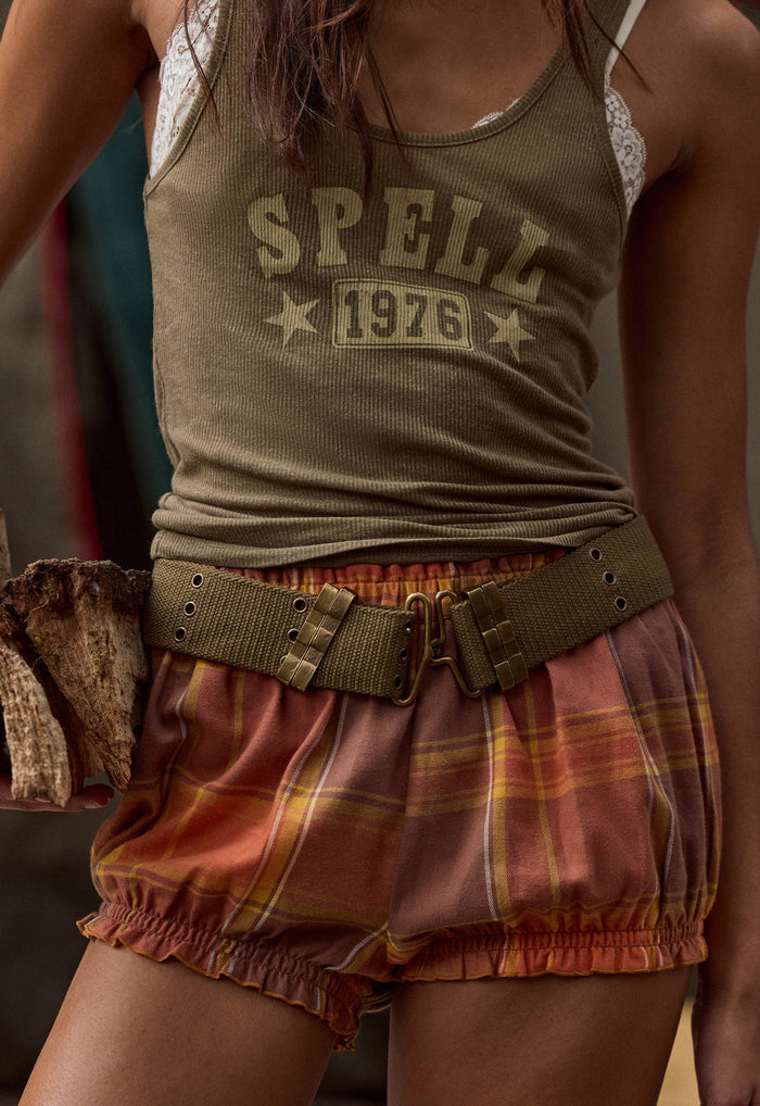 A close-up of a person wearing an olive green ribbed tank top with 'SPELL 1976' printed on the front, paired with rust-orange plaid bloomers and a wide olive belt.