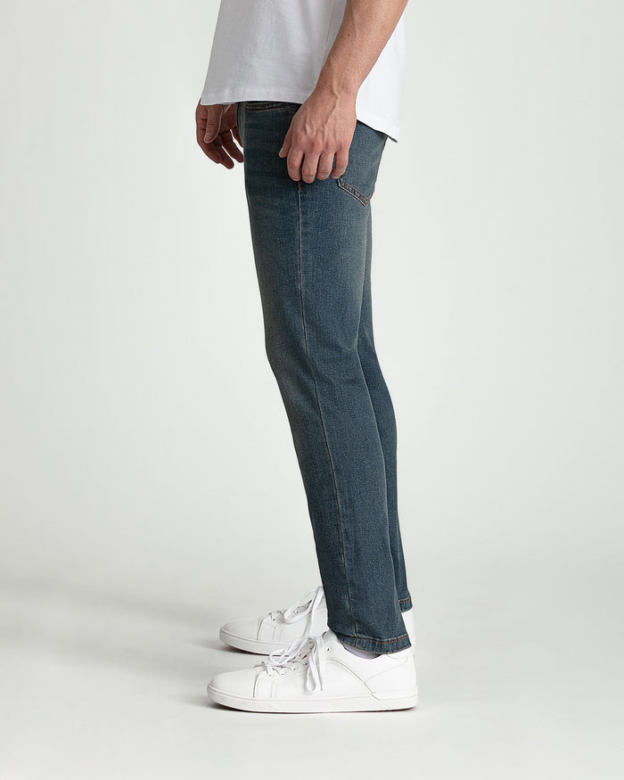Man wearing tinted blue skinny fit jeans with white sneakers and light gray shirt.