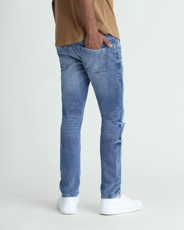 Medium blue skinny fit jeans with ripped details on thighs, shown on model wearing tan shirt and white sneakers.