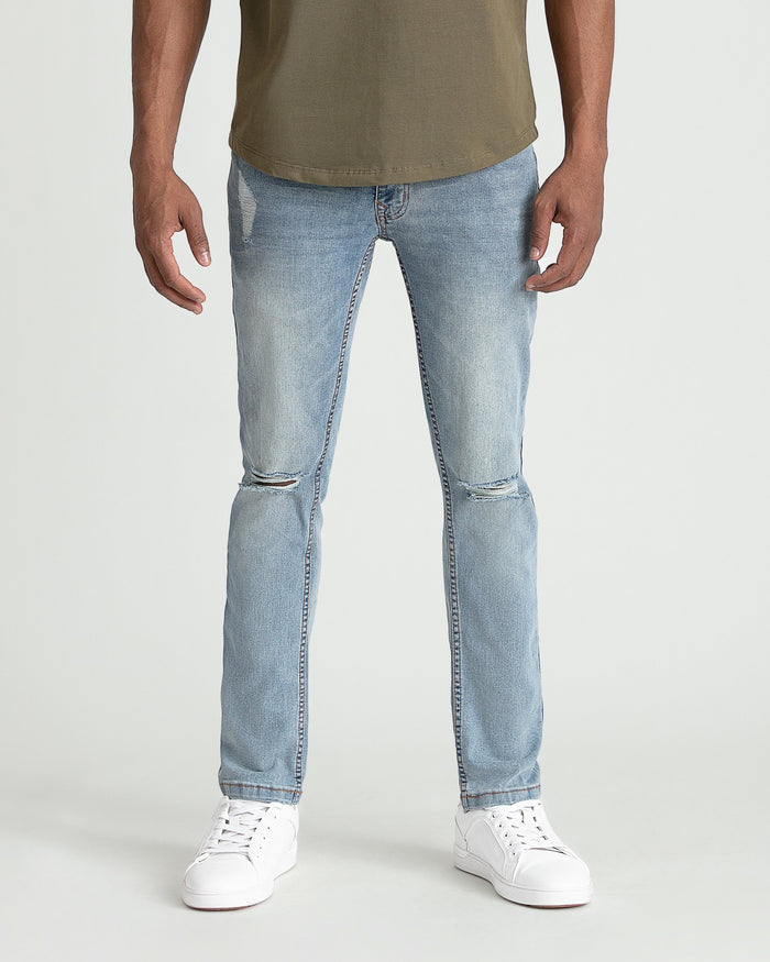 Light wash skinny fit jeans with ripped knees on male model wearing olive t-shirt and white sneakers.