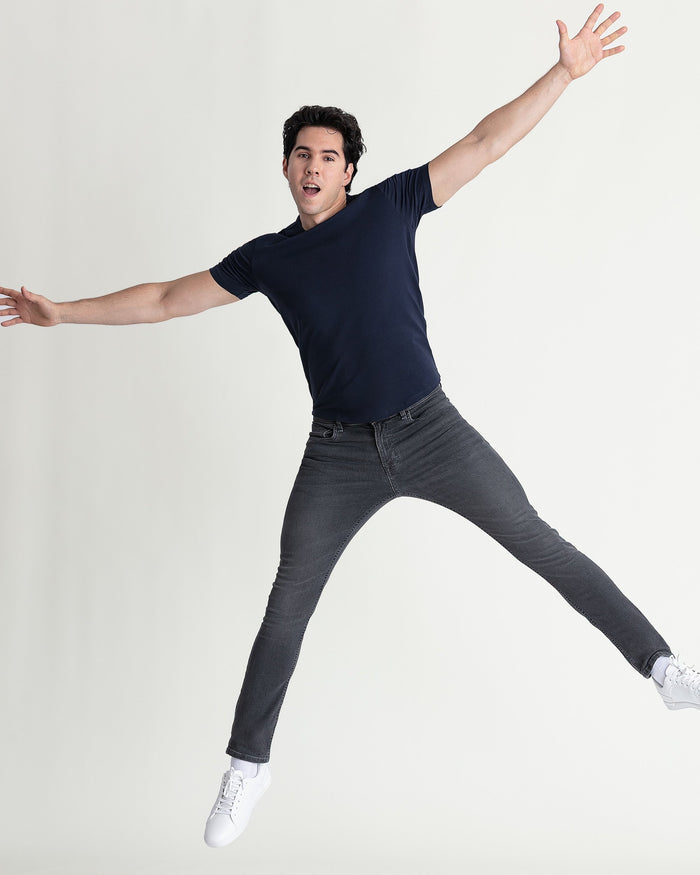 Man wearing dark navy t-shirt and grey skinny fit jeans with white sneakers, jumping with arms spread wide.