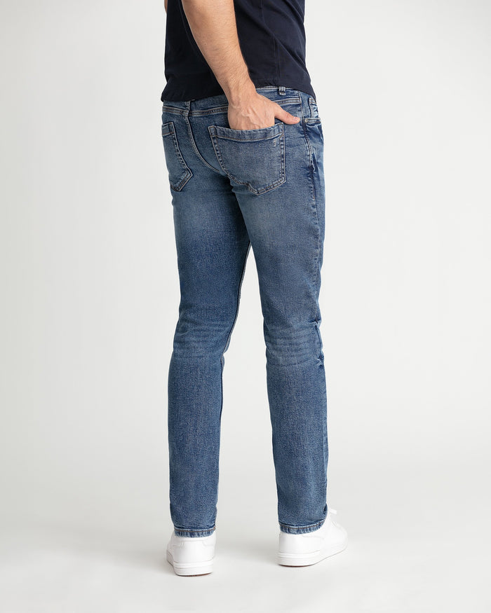Medium blue skinny fit jeans with five-pocket styling, shown on model wearing navy shirt and white sneakers.