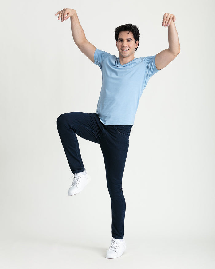Man wearing light blue t-shirt and deep blue skinny fit jeans with white sneakers, arms raised.