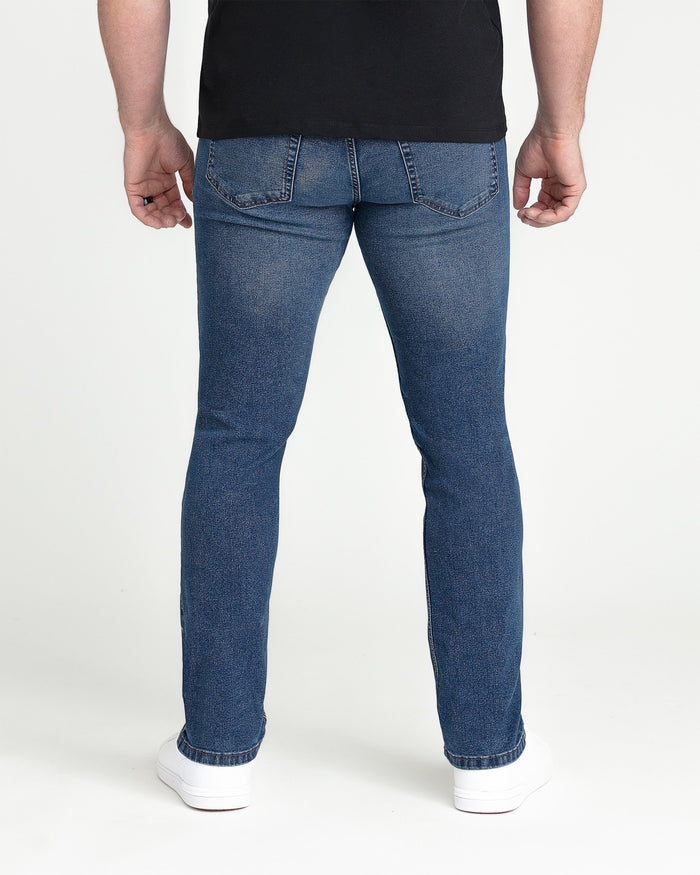 Man wearing medium blue slim fit jeans with black shirt and white sneakers, back view.