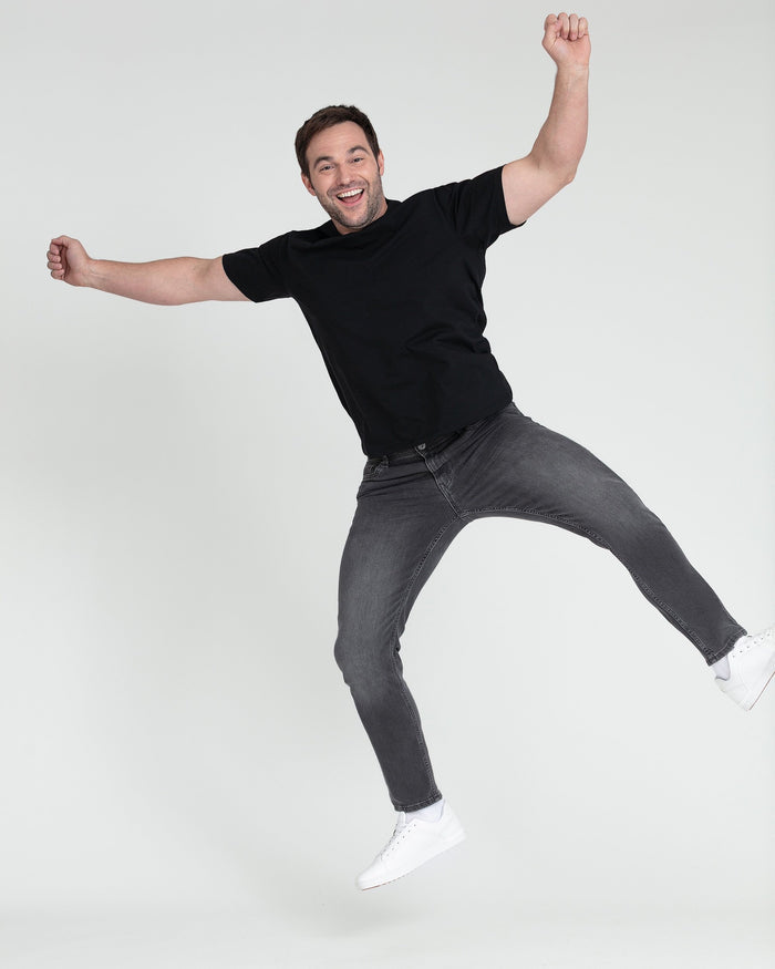 Man wearing black t-shirt and grey slim fit jeans, jumping with arms raised.