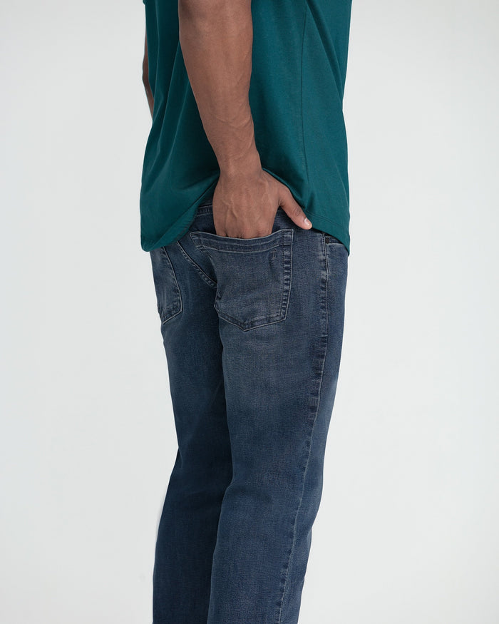 Man wearing midnight blue slim fit shirt tucked into dark blue jeans with hand in pocket.