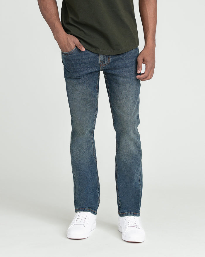 Men's slim fit jeans in medium blue denim with white sneakers and olive green t-shirt.