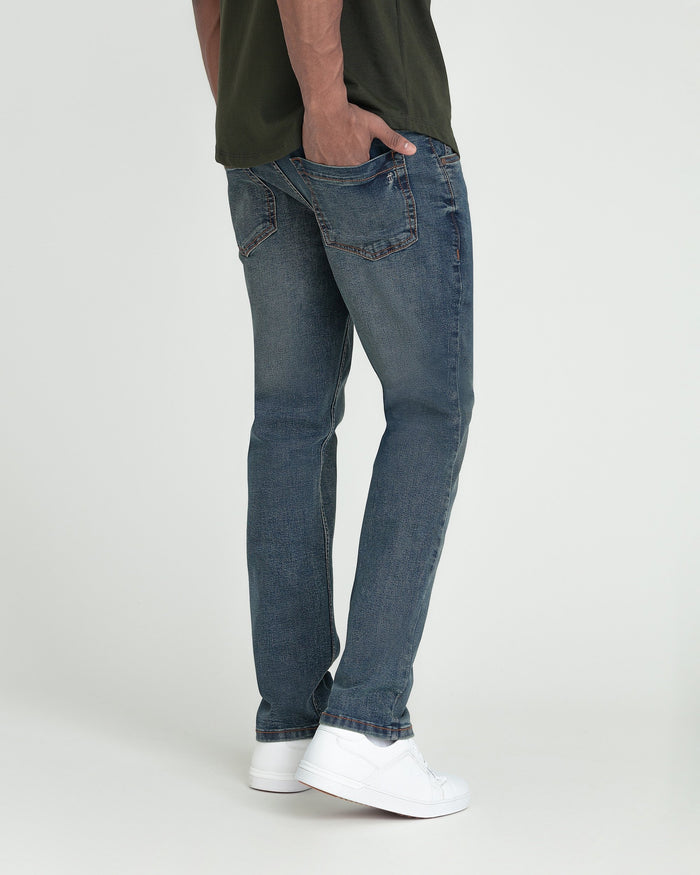 Slim fit medium-wash denim jeans with straight leg, worn with olive shirt and white sneakers.