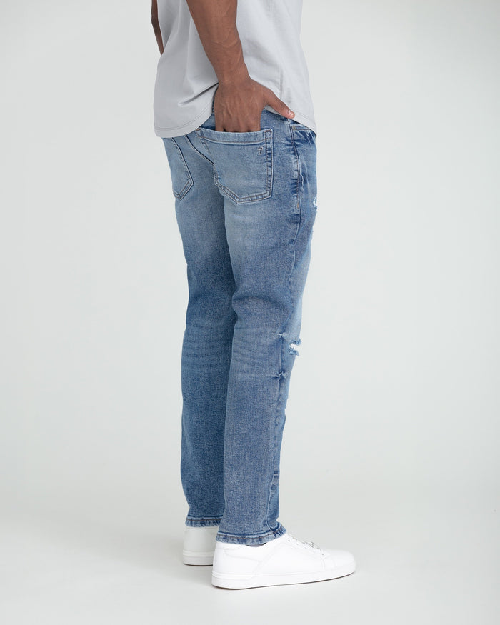 Medium blue slim fit jeans with ripped detail on thighs, shown on model wearing white sneakers.