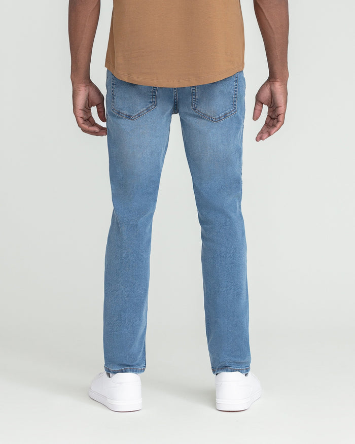 Light indigo slim fit jeans with five-pocket styling shown from back, paired with tan shirt and white sneakers.