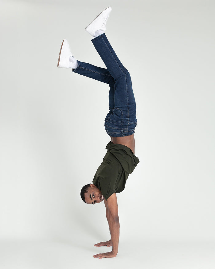 Person doing a handstand wearing dark blue slim fit jeans, olive jacket, and white sneakers.