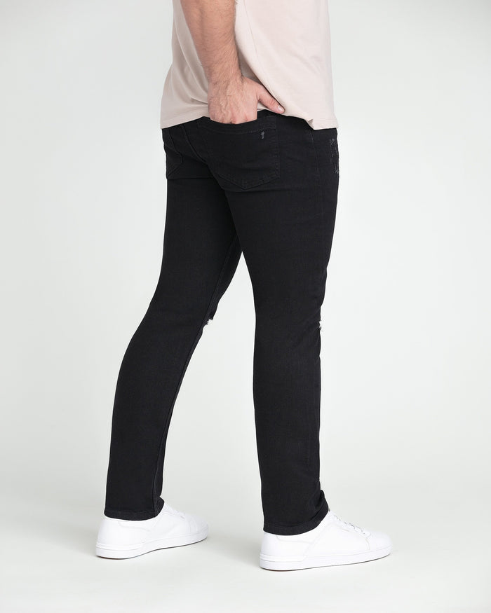 Black slim fit jeans with ripped thigh detail on model wearing white sneakers.