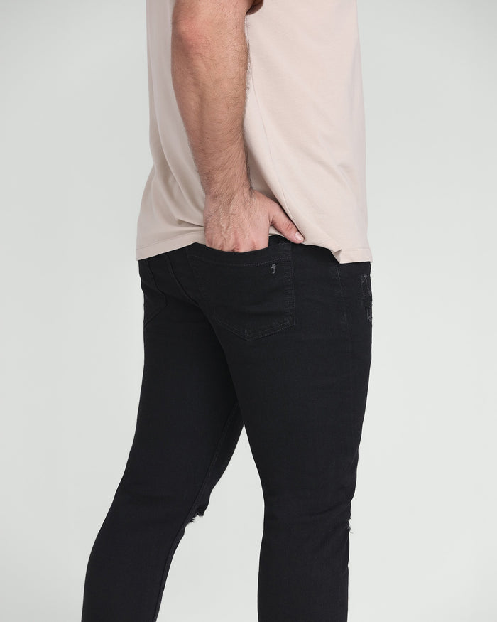 Black slim fit jeans with ripped details at the knees, shown on model wearing cream shirt.